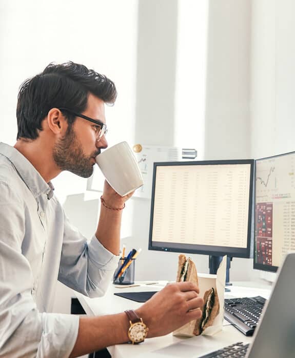 Man working at desk with multiple monitors and eating sandwich, enjoying coffee.