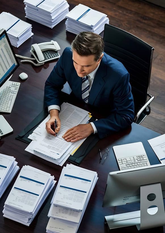 High-angle view of businessman reviewing documents at cluttered desk with papers and office equipment.